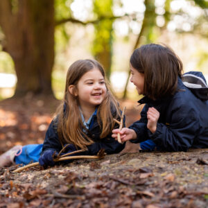 Girls laying on the muddy floor in forest school, discussing their nature finds